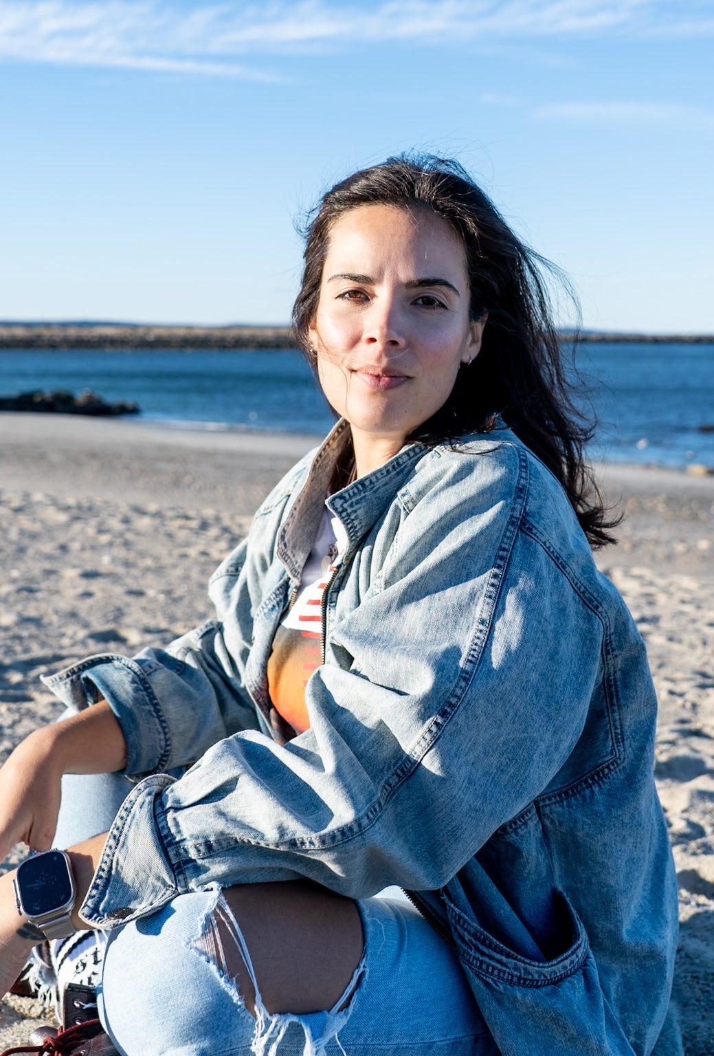 Erika Fernandes-Rusan sitting on the beach in a denim jacket