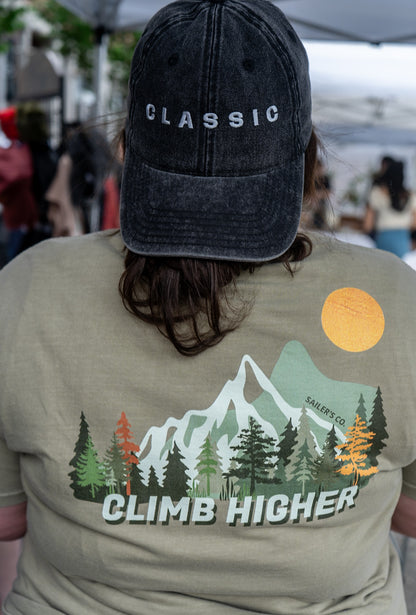 Back view of a person wearing a black Classic embroidered cap and an olive Climb Higher mountain graphic tee