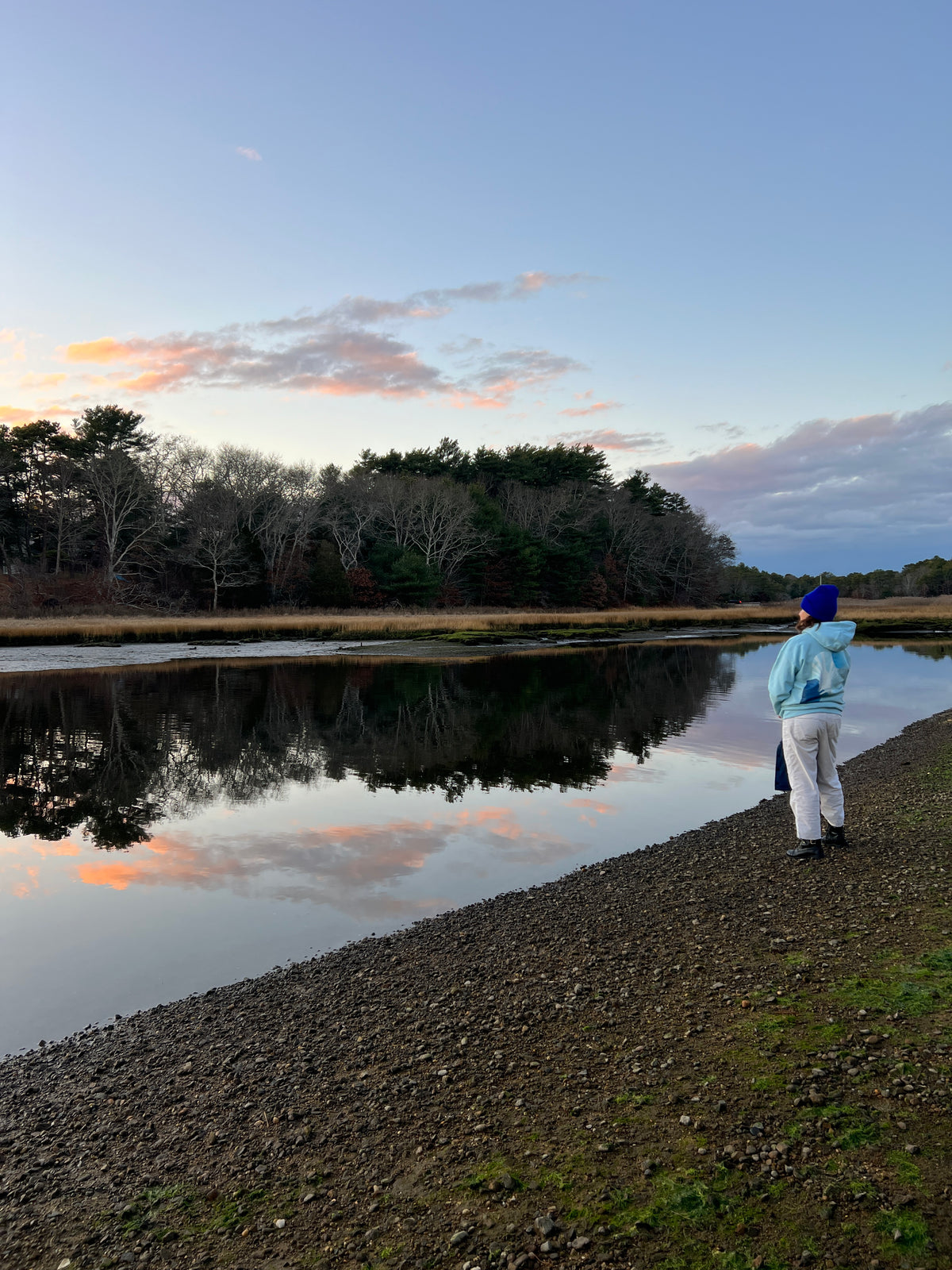 Person in light blue hoodie and blue beanie standing on a pebbly shore at sunset, gazing at calm reflective water.