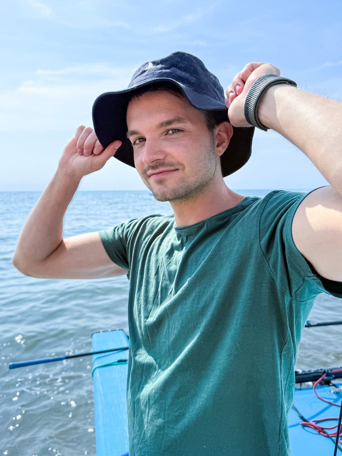 Young man in a black bucket hat and green t-shirt adjusting his hat on a catamaran