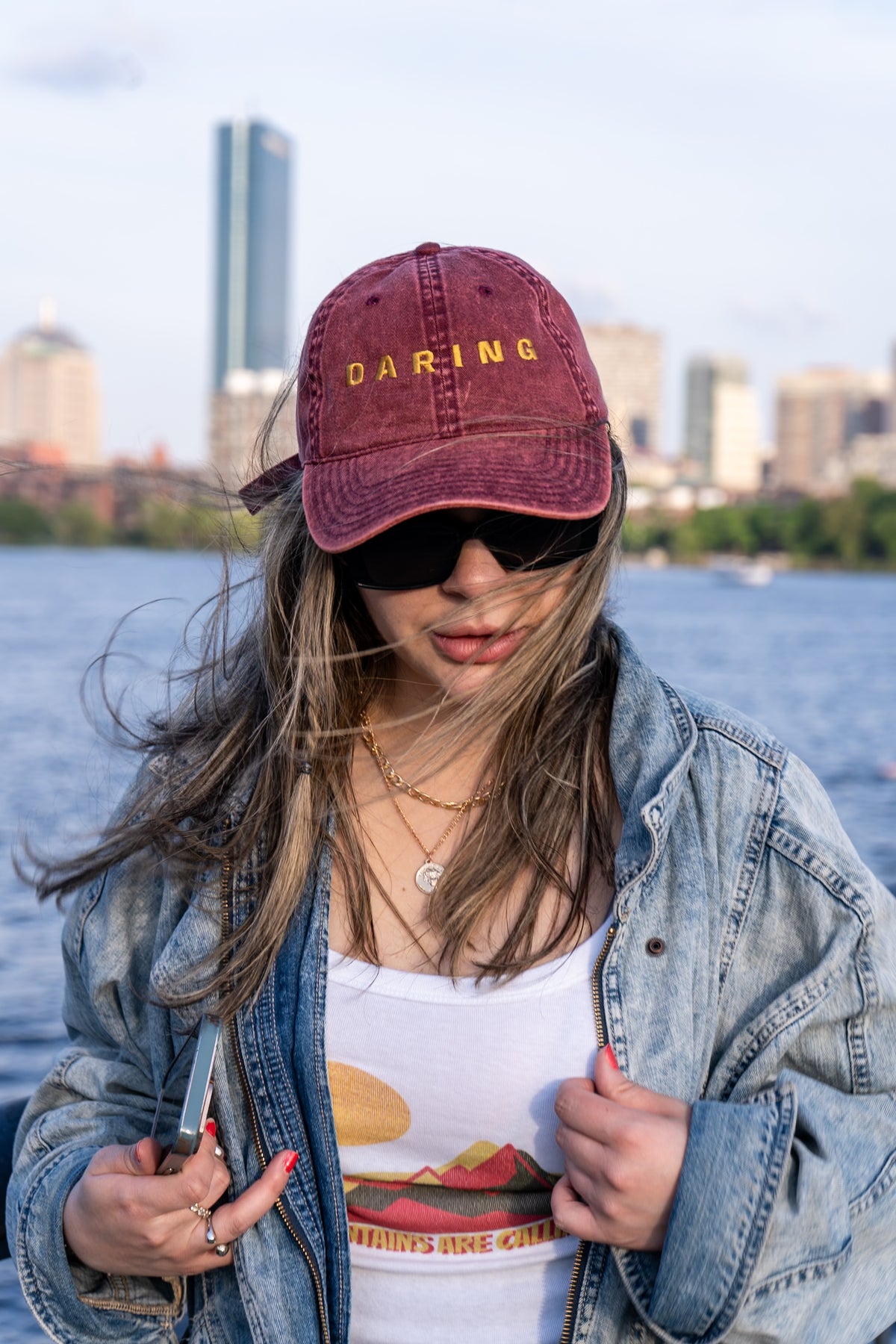 Person wearing a maroon cap with 'DARING' text, sunglasses, and denim jacket by a waterfront with city skyline.