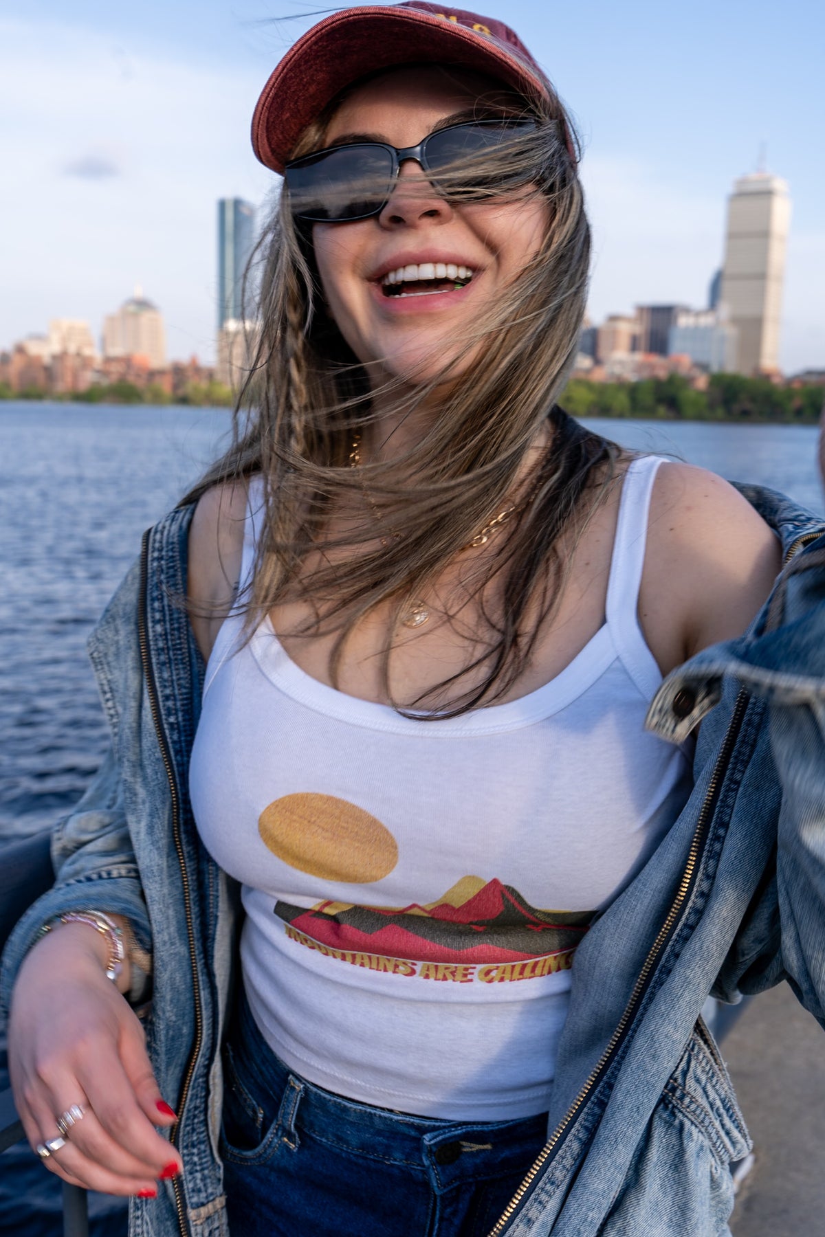 Woman wearing a white tank top with a graphic design, sunglasses, and a red cap by a waterfront with city skyline.